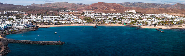 Aerial view of a crescent bay in Lanzarote, Canary Islands, with white resorts, lava breakwaters, jetties, and a lighthouse marker. Boats and a sailboat rest at midday. © Aerial Film Studio