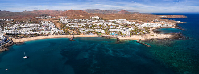 Aerial view of Costa Teguise, Lanzarote, shows white buildings, golden coves, turquoise shallows, curved breakwaters, a marina with a lone catamaran, and ochre volcanic hills. © Aerial Film Studio