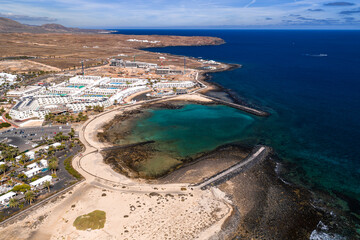 Aerial view shows a turquoise cove, crescent sand beach, and volcanic rock breakwaters on Lanzarote, Canary Islands. Whitewashed resorts, pools, palms, and russet lava fields. © Aerial Film Studio