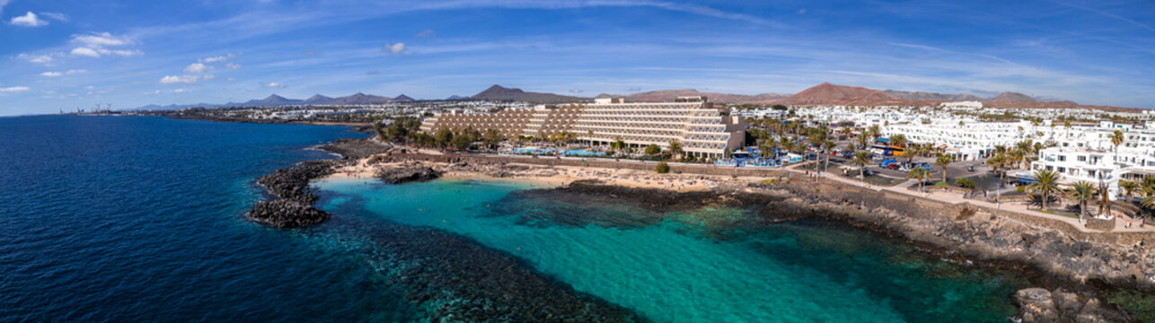 Aerial view of Puerto del Carmen in Lanzarote shows a volcanic shore, a sandy cove, a terraced beachfront hotel, palm lined promenade, and rust red cones near Arrecife.