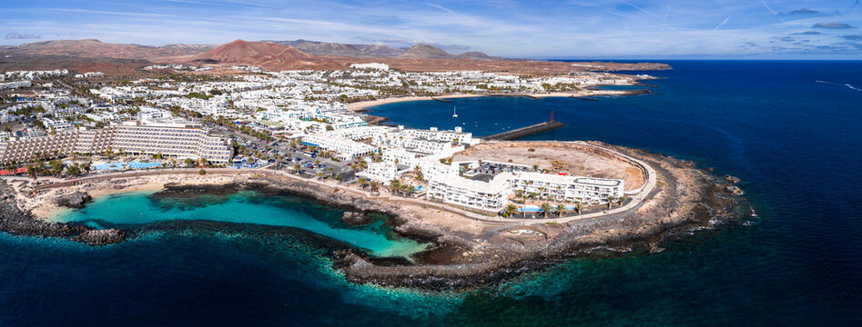 Aerial view of Costa Teguise in Lanzarote shows whitewashed hotels, turquoise coves, rocky promontory, arid volcanic hills, small harbor with sailboats, and midday light.