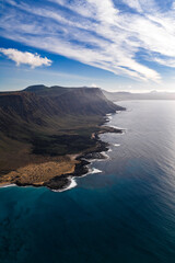 Fototapeta premium Aerial midday view shows Famara cliffs on Lanzarote facing La Graciosa, layered lava fields, dark rocky outcrops, and surf lines along turquoise Atlantic waters under cirrus clouds.