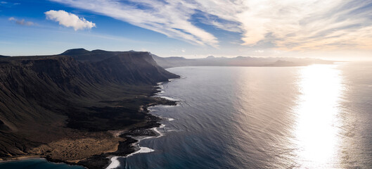 Fototapeta premium Aerial view shows Famara cliffs on Lanzarote, the strait to La Graciosa, dark lava fields, black rock shore, distant volcanic cones, and wispy high clouds at late afternoon.