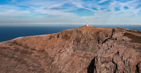 Aerial view shows Mirador del Rio in northern Lanzarote, a communications mast on the plateau, rust colored volcanic cliffs, ravines, a narrow road, and stone walls at midday.