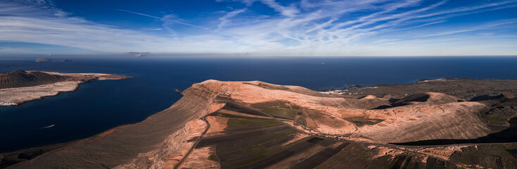 Aerial view of Famara cliffs and La Graciosa near Mirador del Rio, Lanzarote. Terraced black lava fields, La Geria vineyards, road along ridge, whitewashed coastal towns, midday light. © Aerial Film Studio