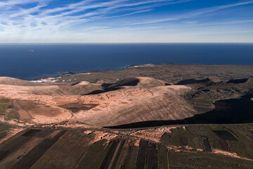 Aerial view of Lanzarote volcanic cones and lava fields leading to the rugged coast near Famara. La Geria plots and a winding road appear, with white buildings by the shore.