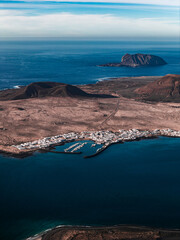 Fototapeta premium Aerial view of a whitewashed village and marina on north Lanzarote, Canary Islands, with cinder cones, lava fields, turquoise shallows, and La Graciosa on the horizon.