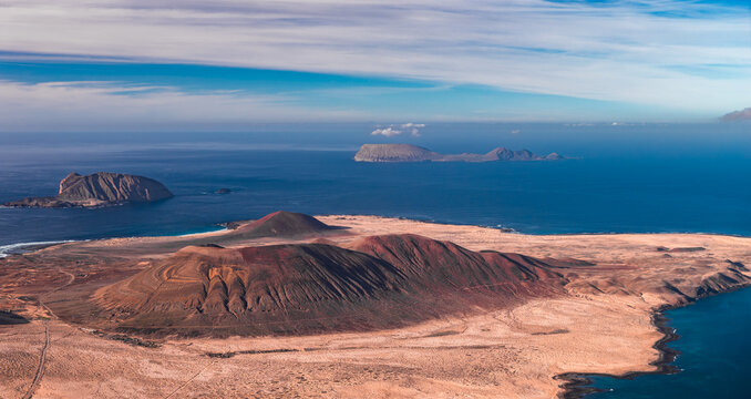 Aerial view of northern Lanzarote shows cinder cones, caldera rims, and lava slopes. La Graciosa and Chinijo islets appear beyond the Mirador del Rio coast under midday light.