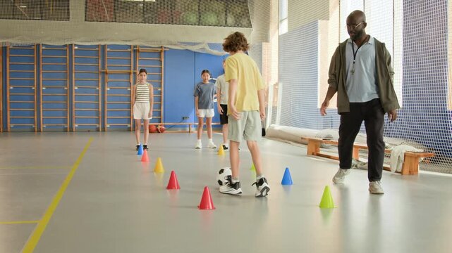 Full length shot of group of children doing soccer drills in PE class with focus on boy leading ball around cones and teacher watching, copy space