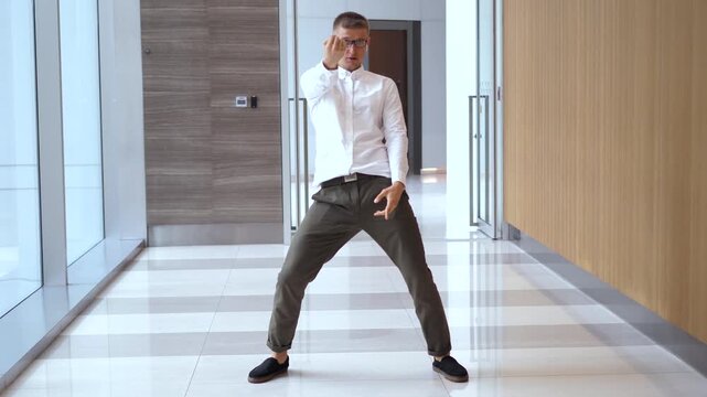 Joyful young professional in glasses and white shirt expressing happiness by dancing enthusiastically and comically in a modern, bright office corridor, celebrating a successful achievement