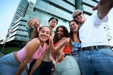 Five cheerful friends take a playful selfie in a modern city, laughing and flashing peace signs by...