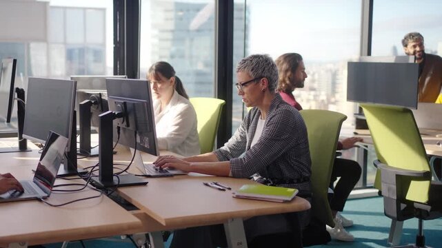 Middle aged professional works at an open plan desk in a bright corporate office, concentrating on tasks at a dual monitor setup with paperwork and notes by the window overlooking the city.