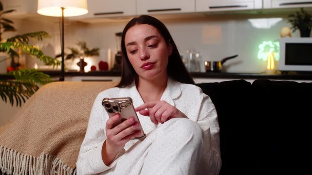 Young woman scrolls news at night home bored tired looks at smartphone sighs stressed unmotivated. Caucasian girl on sofa at evening shows fatigue burnout mood dull scroll stare poor focus wants rest.