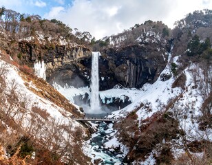 A majestic waterfall cascades down a rocky cliff, partially frozen with snow. People stand on a bridge, appreciating the winter scene