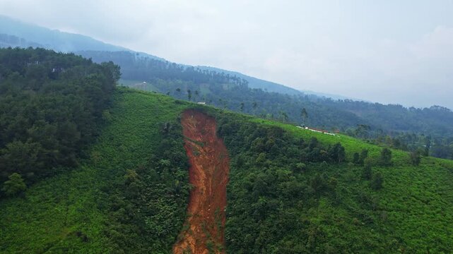 Aerial drone footage of a brown red mudslide in a green hill, with slopes and forests behind, in cloudy weather, in Cipasung, Majalengka regency, Java island, Indonesia