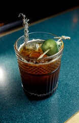 Dark beverage in a clear glass with a pickle garnish on a blue tabletop