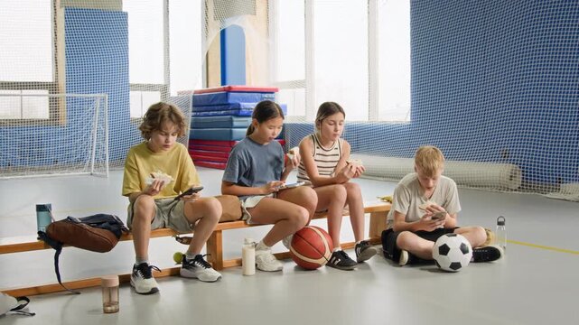 Zoom in shot of group of children sitting on bench in indoor gym after sports practice and eating lunch using phones, copy space