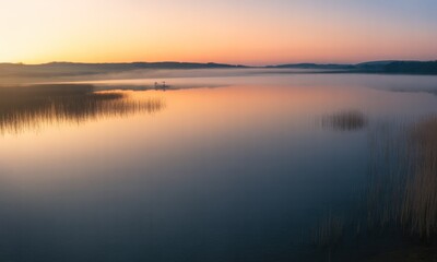 Fototapeta premium Calm lake reflects sunrise hues, light mist shrouds distant shore, reeds in foreground