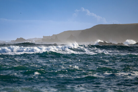 A rough sea of high waves, mist, whitecaps, foam, intense, rapid, strong wings moving turbulent green water onshore. There's a rocky coastline with a bright blue sky in the distance. The choppy waves 