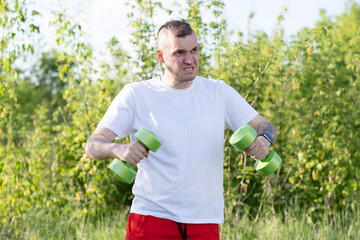 A man exercises outdoors in a park, lifting green dumbbells under a clear sky during daytime