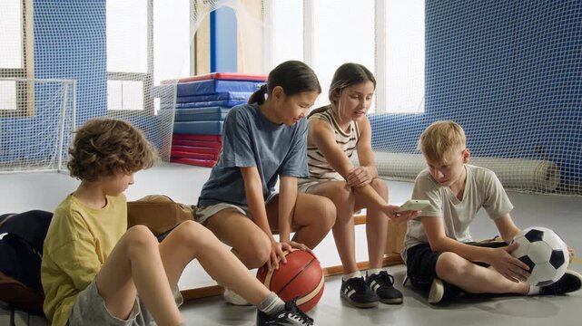Full length shot of group of children sitting on bench and chatting during break looking at smartphone screen and playing with sports balls