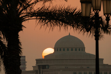 The sun sets behind a dome structure while a palm tree stands in the foreground. The scene captures...