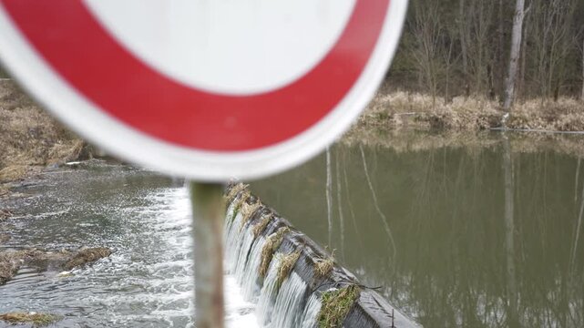 Slow pan showcasing a dammed river.  Water cascades over a barrier.  Vegetation lines the banks.  A no-entry sign appears.  Calm, natural scenery.  Peaceful flow