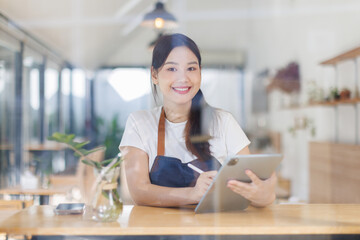 Portrait of happy asian woman using digital tablet in her store. 
Successful sme small business owner in casual wearing blue apron at entrance cafe.