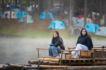 asian family by mother daughter tourist wearing black coat and relax travel on bamboo rafting in lake with morning fog mist on water surface and camping tent at Pang Ung Reservoir in winter thailand