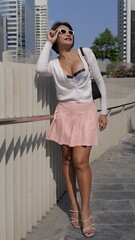 Fototapeta premium Stylish woman in a pink pleated skirt and white strappy sandals stands against a modern urban backdrop