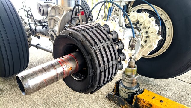 Close-up view of a wide-body aircraft landing gear raised on hydraulic jacks, with one wheel removed to reveal the brake assembly, showcasing aviation engineering and heavy aircraft maintenance