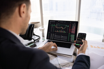 Professional trader working at laptop, using stock market exchange application on smartphone for managing assets, investment. Over shoulder view of displays with financial charts, diagrams