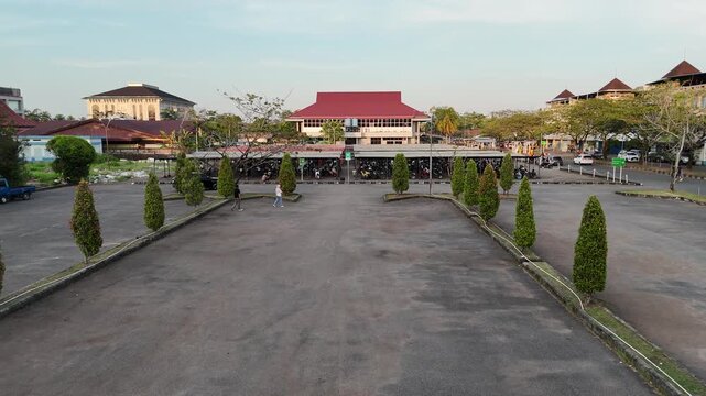 A large concrete parking lot at dusk with neat rows of green shrubs along its edges, rows of motorcycles under a long roof in front of a large red-roofed building with a gabled roof, trees, and passin