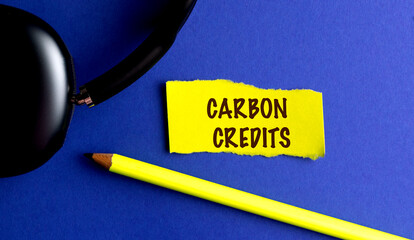 Carbon credits concept written on yellow torn paper with pencil and headphones on blue background