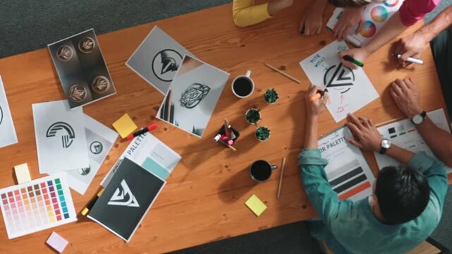 Top view of designer writing and designing logo graphic at meeting. Closeup of business man working on colored palettes and writing idea on sticky notes on table with equipment and paper. Symposium.