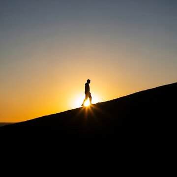 Man walking uphill at sunset, silhouette against vibrant orange sky, overcoming challenge
