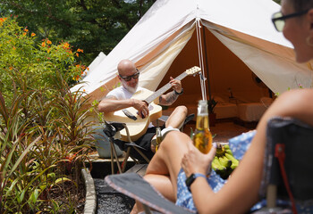 A man and woman is joyfully drinking beer and playing guitar near a cozy tent at glamping