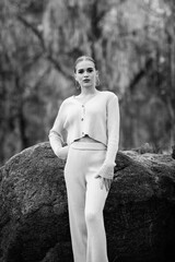 Young woman in a black and white photograph wearing a stylish jacket and wide-legged pants poses confidently in a forested area with fallen leaves on the ground