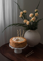 Birthday sponge cake with cream and bouquet of flowers on the wooden table in the living room