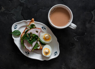 Delicious grilled breakfast - bread toast with cream cheese, spinach, ham, pumpkin seeds, boiled egg and coffee with milk on a dark background, top view