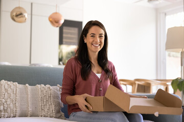 Latina woman sit on sofa with cardboard delivery package with excitement, looks at camera. Portrait of satisfied online shopping client enjoy fast delivery, received gift and moment of parcel unboxing