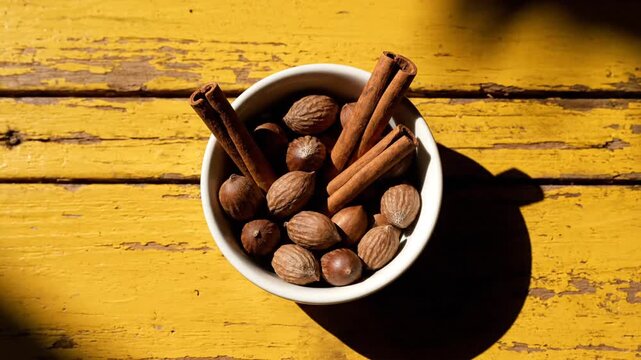 A static, top-down shot showcases a white bowl filled with whole nutmegs and cinnamon sticks, resting on a rustic, brightly-lit yellow wooden surface, emphasizing deep shadows and texture.