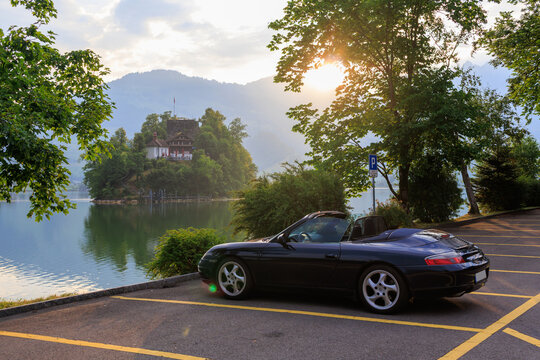Lauerz, Switzerland - July 5, 2025: Blue Porsche 911 convertible and ruined castle Burgruine Schwanau, chapel and restaurant on island Schwanau in Lake Lauerz, located in the canton of Schwyz
