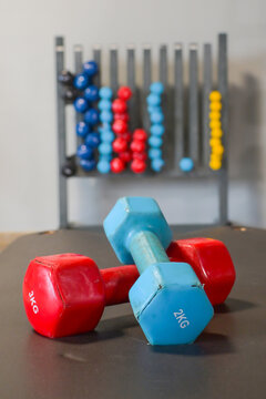 Two colored dumbbells, a 3kg red and a 2kg blue, resting on a dark mat in a gym with blurred weight rack in the background, signifying fitness, strength, and endurance