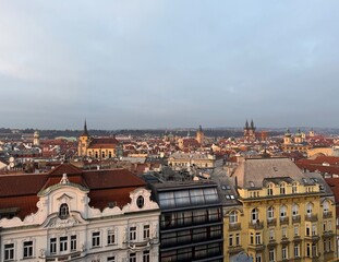 Obraz premium View over a dense cityscape with red-tiled rooftops, historic buildings, and multiple church towers in the distance. Foreground shows ornate facades and multi-story residential buildings with windows.