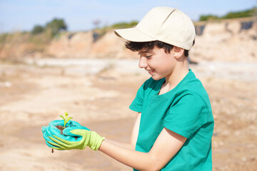 Smiling boy wearing a cap and green gloves holding a small plant in his hands before planting it in the ground.