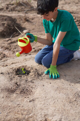 Smiling child wearing green gloves holding a small watering can planting a small plant in the soil.