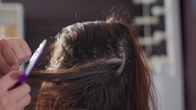 caucasian colorist applying root touch up in top sections, steady brush strokes and parted layers, even coverage and neat parting, studio shelf of color products in background, professional technique