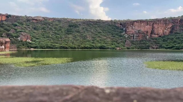 A mesmerizing time-lapse of Agastya Lake, featuring calm reflective waters with the towering red sandstone mountains in the background, showcasing the tranquil and majestic beauty of Badami.