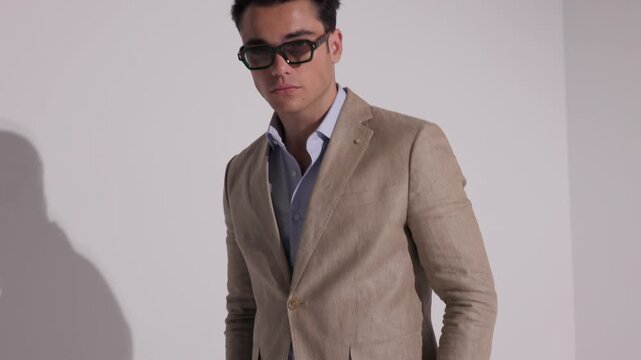 Stylish man in beige suit and sunglasses turning to camera, grabbing jacket lapels and posing arrogantly on white studio background.